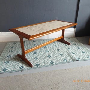 May include: A mid-century modern coffee table with a tiled top. The table has a light wood frame and a white tiled top with a floral pattern. The table is sitting on a green and white patterned rug.