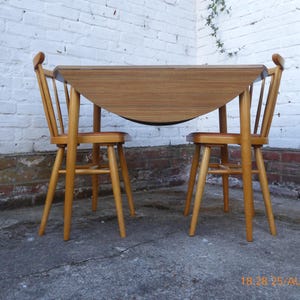 May include: A wooden drop-leaf table with a smooth, light brown grain finish, paired with two matching wooden chairs featuring spindle backs and brown upholstered seats. The table has one side folded down, revealing the underside construction.