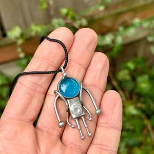 May include: A silver pendant necklace with a blue glass circle and a stylized figure with arms and legs. The pendant is on a black cord.