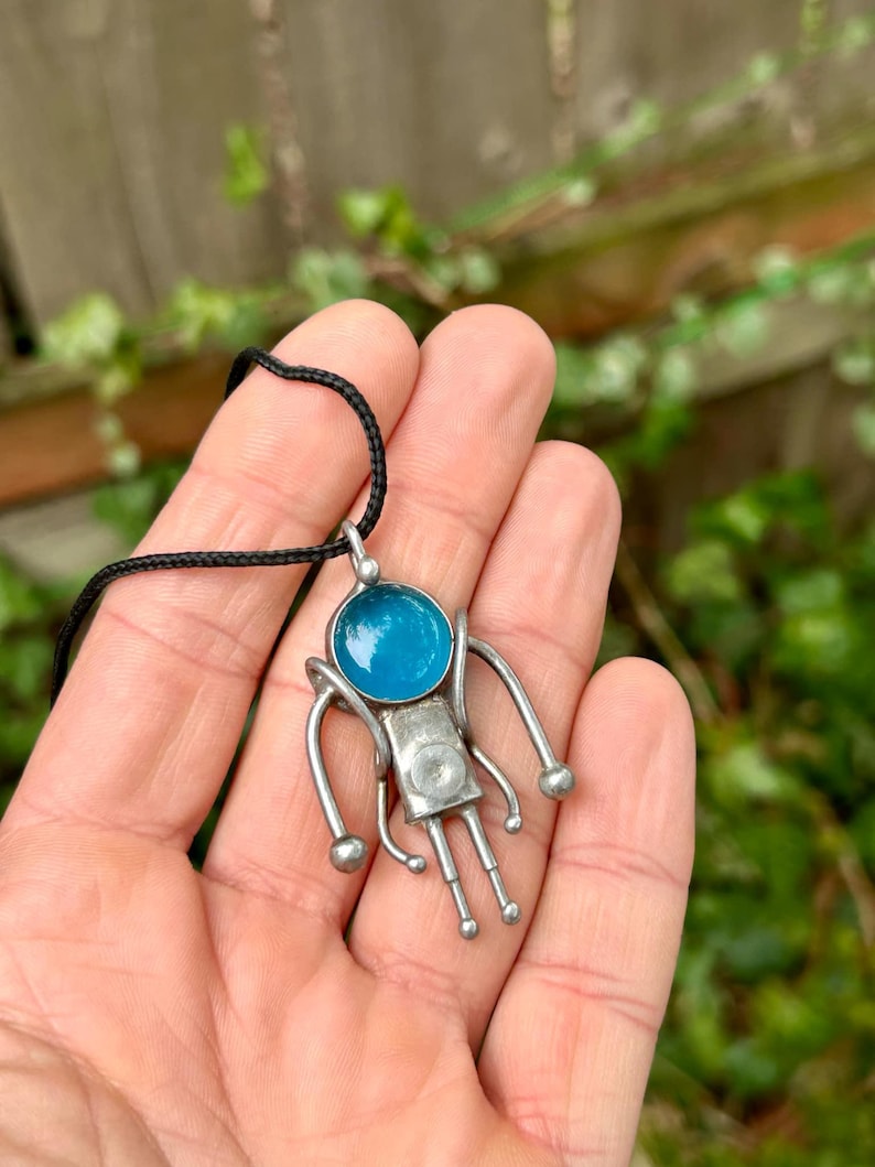 May include: A silver pendant necklace with a blue glass circle and a stylized figure with arms and legs. The pendant is on a black cord.