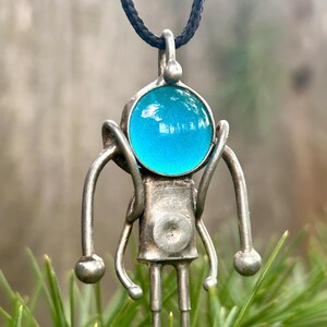 May include: A silver robot pendant with a blue glass head, hanging from a black cord. The robot has long arms and legs, and a round body.
