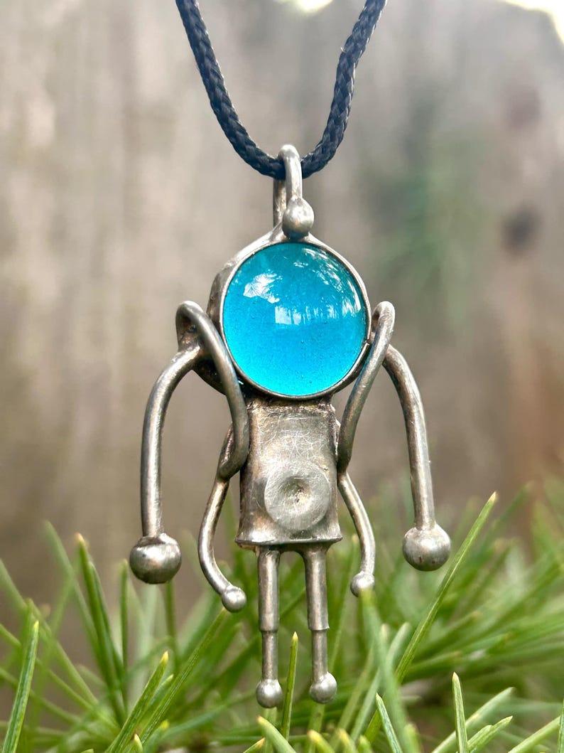 May include: A silver robot pendant with a blue glass head, hanging from a black cord. The robot has long arms and legs, and a round body.