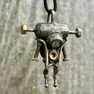 May include: A steampunk robot pendant necklace. The metal pendant has a tarnished, aged finish, with a cylindrical body, bolt-like arms, and a circular head with a symbol. It hangs from a black cord. The design evokes a retro-futuristic aesthetic.