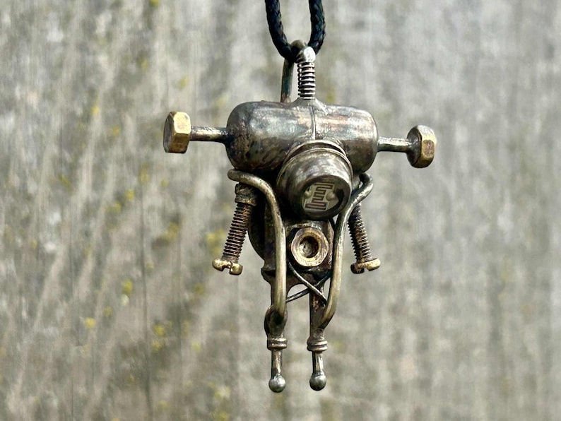 May include: A steampunk robot pendant necklace. The metal pendant has a tarnished, aged finish, with a cylindrical body, bolt-like arms, and a circular head with a symbol. It hangs from a black cord. The design evokes a retro-futuristic aesthetic.