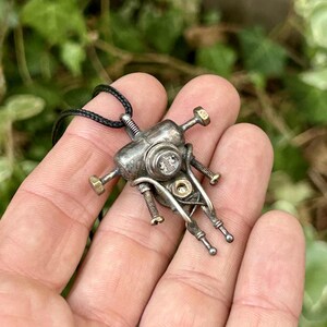 May include: A silver pendant necklace with a distinctive mechanical design. The pendant incorporates bolts, screws, and small metal components, giving it a steampunk appearance. The necklace has a black cord.