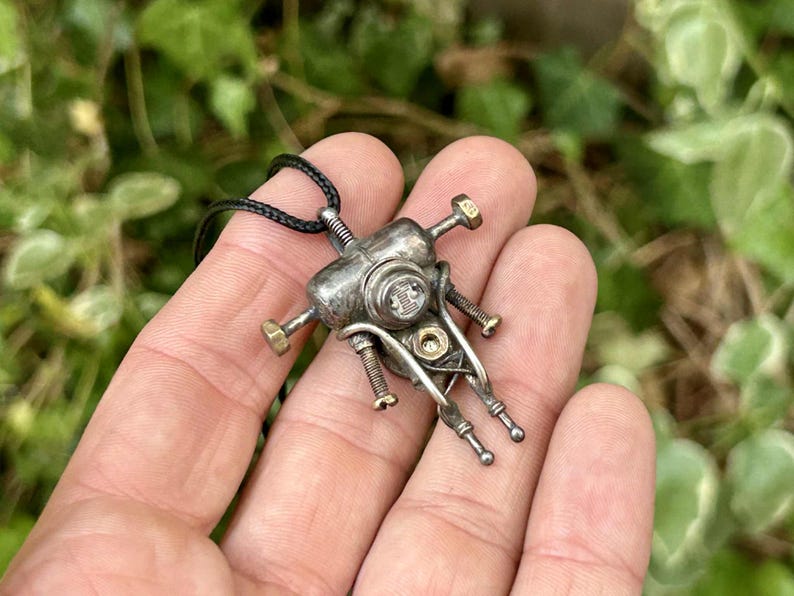 May include: A silver pendant necklace with a distinctive mechanical design. The pendant incorporates bolts, screws, and small metal components, giving it a steampunk appearance. The necklace has a black cord.