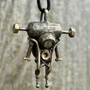 May include: A metal pendant necklace in a steampunk robot design. The pendant has a cylindrical body, arms, and legs, with brass bolts and screws. The robot is suspended from a black cord. The pendant's dimensions are approximately 5 cm in height.
