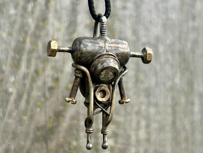 May include: A metal pendant necklace in a steampunk robot design. The pendant has a cylindrical body, arms, and legs, with brass bolts and screws. The robot is suspended from a black cord. The pendant's dimensions are approximately 5 cm in height.