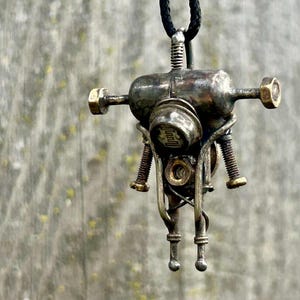 May include: A steampunk pendant necklace with a metallic robot-like design. The pendant has an aged, industrial look, incorporating bolts, screws, and a central cylindrical element. It is suspended from a black cord.