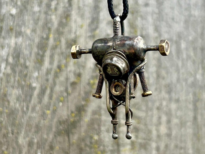 May include: A steampunk pendant necklace with a metallic robot-like design. The pendant has an aged, industrial look, incorporating bolts, screws, and a central cylindrical element. It is suspended from a black cord.