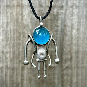 May include: A silver pendant necklace with a blue glass orb and a stylized figure design.