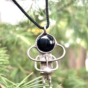 May include: A silver pendant necklace with a black gemstone and a stylized figure design. The pendant is hanging from a black cord.