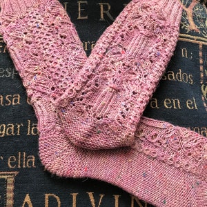 Op de afbeelding: Een paar roze gebreide sokken met een kantpatroon. De sokken hebben een gespikkelde uitstraling met kleine vlekjes kleur.