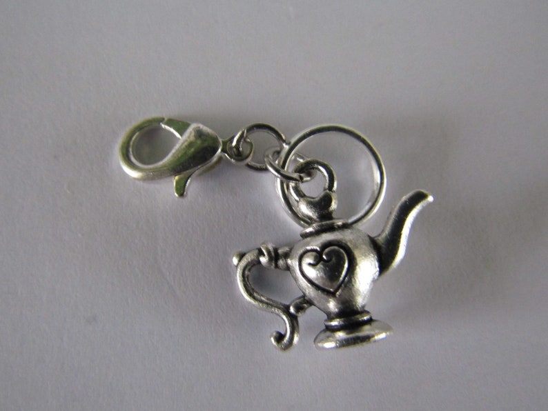 May include: Silver charm in the shape of a teapot with a heart design. The charm has a lobster clasp for attaching to a bracelet or necklace.