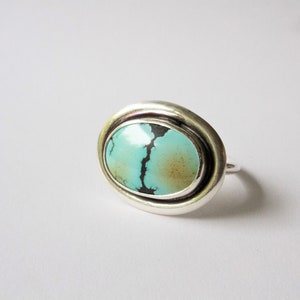 May include: A silver ring featuring a large, oval-shaped turquoise stone. The stone is light blue with a prominent dark brown matrix vein running vertically through the center and a soft brown patch on the right side, set in a smooth silver bezel.