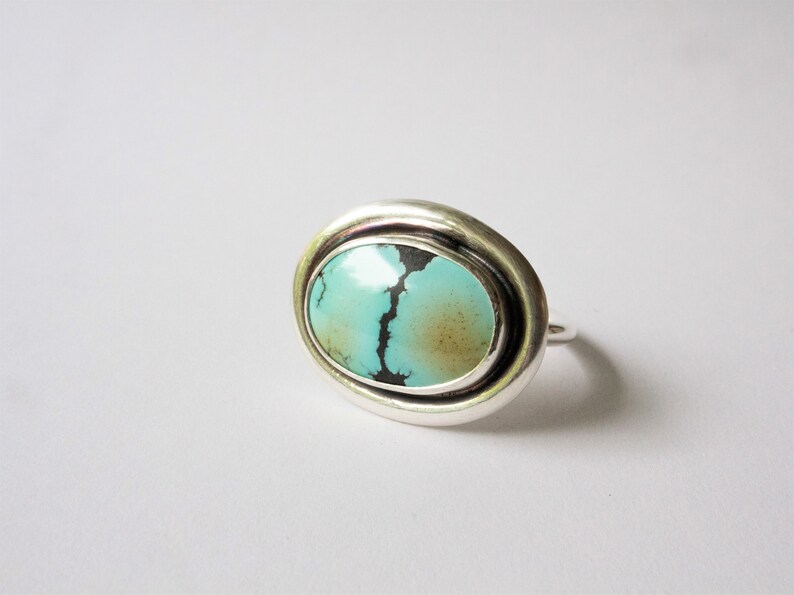 May include: A silver ring featuring a large, oval-shaped turquoise stone. The stone is light blue with a prominent dark brown matrix vein running vertically through the center and a soft brown patch on the right side, set in a smooth silver bezel.