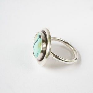 May include: A silver ring featuring a round, polished turquoise stone with dark matrix veining. The stone is set within a smooth, circular silver bezel that sits atop a simple, rounded silver band, showcasing a clean and reflective metallic finish.