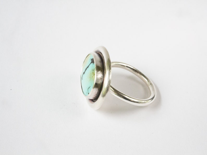 May include: A silver ring featuring a round, polished turquoise stone with dark matrix veining. The stone is set within a smooth, circular silver bezel that sits atop a simple, rounded silver band, showcasing a clean and reflective metallic finish.