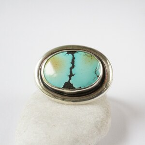 May include: An oval ring featuring a polished turquoise stone with a prominent dark matrix pattern. The stone is set in a smooth, wide silver band that encircles the oval shape, creating a clean and reflective metallic border around the gem.