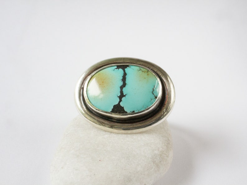 May include: An oval ring featuring a polished turquoise stone with a prominent dark matrix pattern. The stone is set in a smooth, wide silver band that encircles the oval shape, creating a clean and reflective metallic border around the gem.