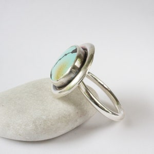 May include: A silver ring featuring a round, light blue turquoise stone with dark matrix veining and a subtle golden-brown patch. The stone is set in a smooth, polished silver bezel, mounted on a simple, rounded silver band with a polished finish.