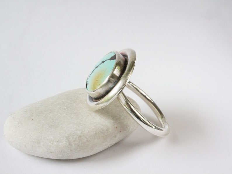 May include: A silver ring featuring a round, light blue turquoise stone with dark matrix veining and a subtle golden-brown patch. The stone is set in a smooth, polished silver bezel, mounted on a simple, rounded silver band with a polished finish.