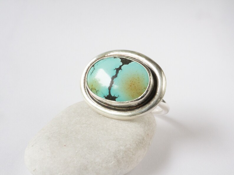 May include: A silver ring featuring an oval-shaped turquoise stone with a smooth, polished surface. The stone displays a vibrant cyan color with a prominent dark brown matrix vein running through the center and a soft golden-brown patch.