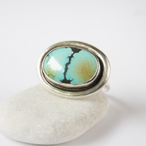 May include: A silver ring featuring a large, oval-shaped turquoise stone. The stone is light blue with a prominent dark brown matrix vein running through the center and a soft brown patch on the right side, set within a smooth, polished silver bezel.