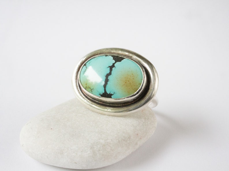 May include: A silver ring featuring a large, oval-shaped turquoise stone. The stone is light blue with a prominent dark brown matrix vein running through the center and a soft brown patch on the right side, set within a smooth, polished silver bezel.