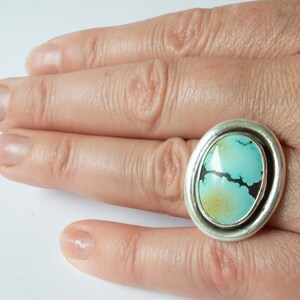 May include: A silver ring featuring a large, oval-shaped turquoise stone with dark matrix veining. The stone displays a gradient of light blue to pale yellow hues, set within a smooth, polished silver bezel and a wide, rounded silver band.