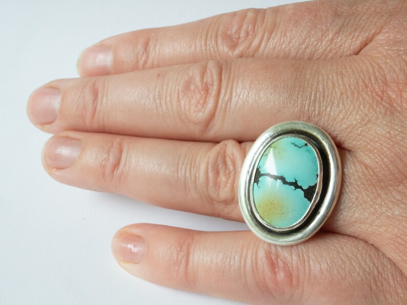 May include: A silver ring featuring a large, oval-shaped turquoise stone with dark matrix veining. The stone displays a gradient of light blue to pale yellow hues, set within a smooth, polished silver bezel and a wide, rounded silver band.