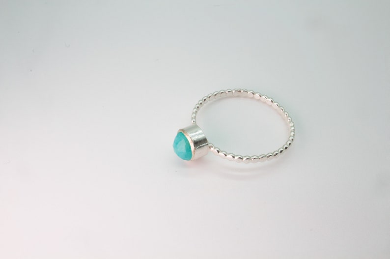 May include: A silver ring with a turquoise gemstone set in a bezel setting. The band is made of small, round beads.