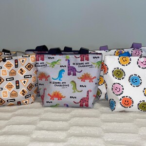 May include: Six different tote bags with various designs. The bags are all white with different colored patterns. One bag has a dinosaur pattern with the text "Be strong and courageous". Another bag has a pattern of road signs. Another bag has a pattern of smiley faces.