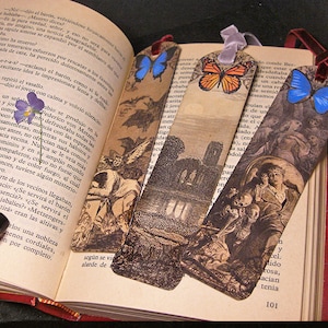 May include: Four decorative bookmarks with vintage illustrations and butterfly accents are displayed open in a book. The bookmarks feature images of landscapes and figures, with ribbon attachments.