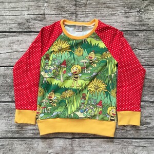Könnte beinhalten: Langarm-Shirt in Rot und Gelb mit weißem Punktmuster an den Ärmeln. Das Shirt zeigt einen bunten Cartoon-Druck mit Bienen, Blumen und anderen Insekten auf einer grünen Wiese.