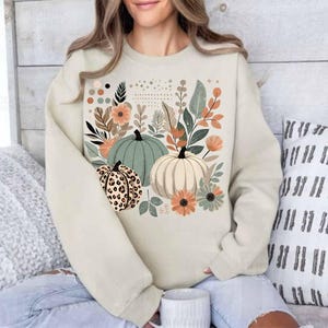 May include: A cream-coloured sweatshirt with a graphic print of pumpkins and autumn foliage. The pumpkins are green, white, and brown with a leopard print pattern.