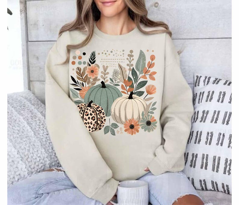May include: A cream-coloured sweatshirt with a graphic print of pumpkins and autumn foliage. The pumpkins are green, white, and brown with a leopard print pattern.