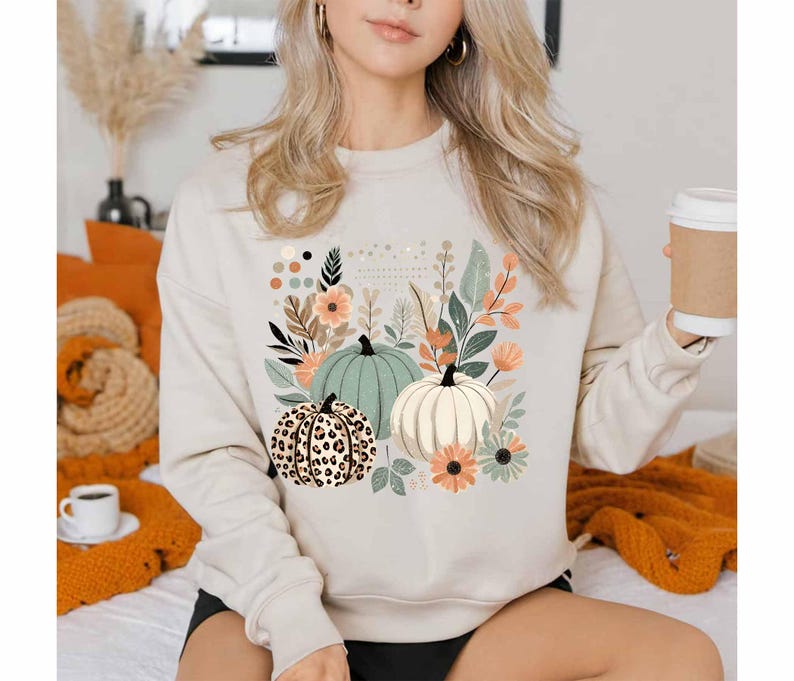 May include: A beige sweatshirt featuring a graphic print of three pumpkins and floral designs. The pumpkins are green, white, and brown with a leopard print pattern.