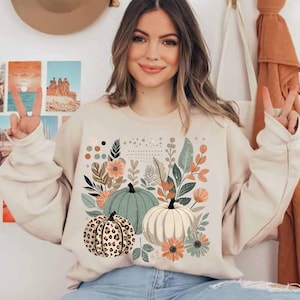 May include: A cream-coloured sweatshirt with a graphic print of three pumpkins and floral designs. The pumpkins are green, white, and brown with a leopard print pattern.