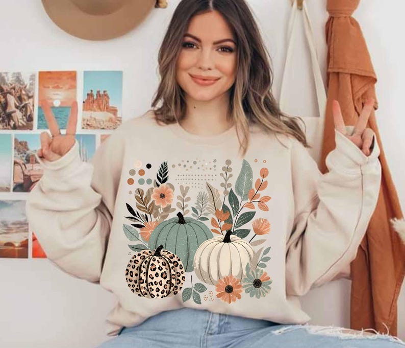 May include: A cream-coloured sweatshirt with a graphic print of three pumpkins and floral designs. The pumpkins are green, white, and brown with a leopard print pattern.