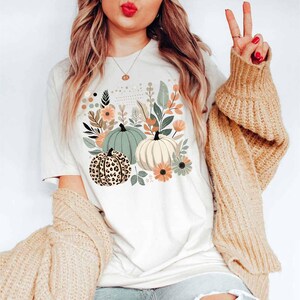 May include: A white t-shirt featuring a graphic of pumpkins and autumn foliage. The pumpkins are green, white, and brown with a leopard print pattern.