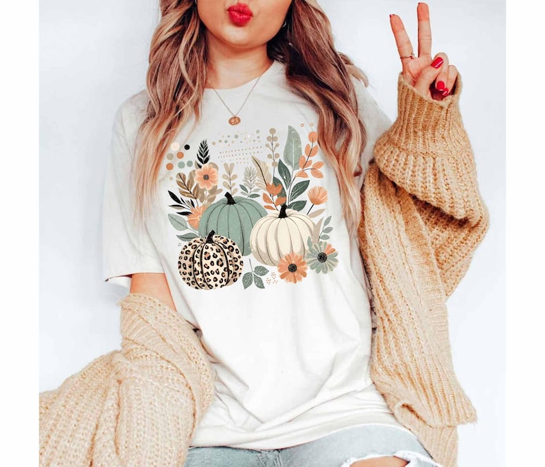 May include: A white t-shirt featuring a graphic of pumpkins and autumn foliage. The pumpkins are green, white, and brown with a leopard print pattern.