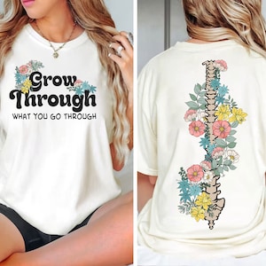 May include: White t-shirt with the text "Grow Through What You Go Through" in black with floral accents. The back of the shirt features a floral design with a spine in the centre.