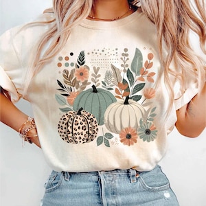 May include: A white t-shirt featuring a graphic print of three pumpkins and floral designs. The pumpkins are green, white, and brown with a leopard print pattern.