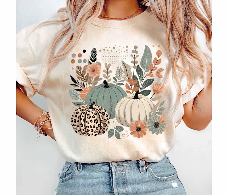 May include: A white t-shirt featuring a graphic print of three pumpkins and floral designs. The pumpkins are green, white, and brown with a leopard print pattern.