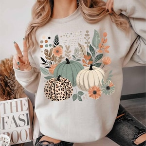 May include: A white sweatshirt featuring a graphic print of three pumpkins and floral designs. The pumpkins are green, white, and brown with a leopard print pattern.