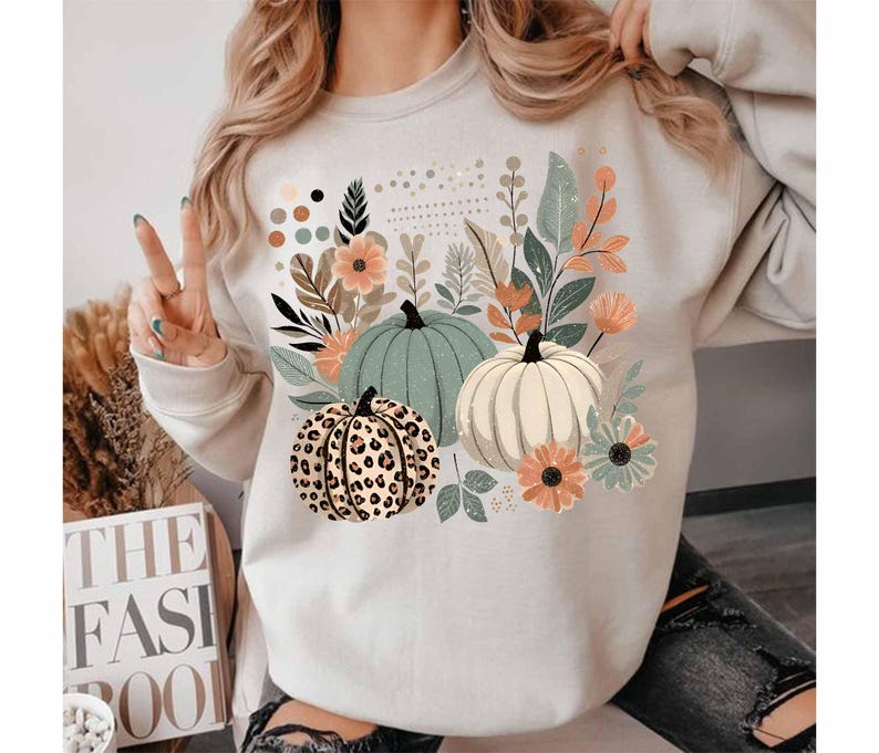 May include: A white sweatshirt featuring a graphic print of three pumpkins and floral designs. The pumpkins are green, white, and brown with a leopard print pattern.