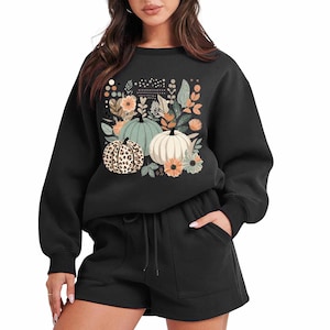 May include: Black sweatshirt with a graphic print of pumpkins and flowers. The sweatshirt has long sleeves and a crew neck. The model is wearing black shorts.