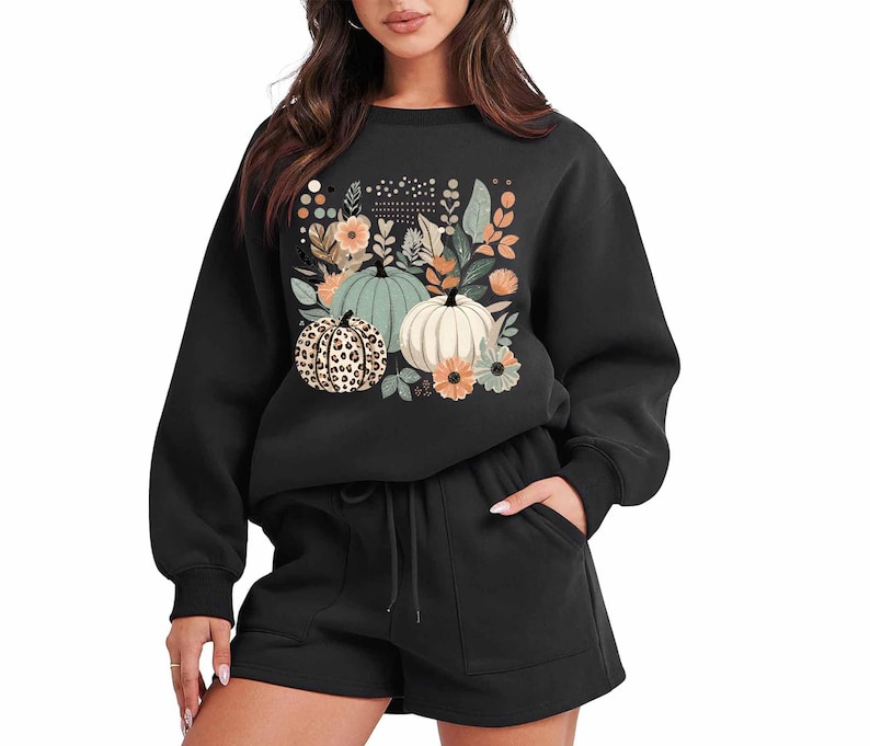 May include: Black sweatshirt with a graphic print of pumpkins and flowers. The sweatshirt has long sleeves and a crew neck. The model is wearing black shorts.