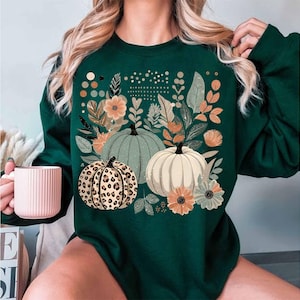 May include: A green sweatshirt featuring a graphic print of pumpkins and flowers. The pumpkins are in various shades of green, white, and brown with a leopard print pattern.